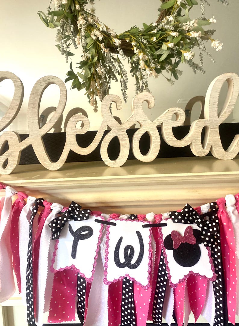 May include: A decorative banner with pink, white, and black polka dot and striped fabric strips. The banner includes three white pennants with the letters "P", "W", and a Minnie Mouse silhouette. A wooden "Blessed" sign and a floral wreath are in the background.