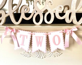 May include: Pink and white banner with the word "TWO" in pink letters. The banner is decorated with pink satin ribbon bows and is hanging from a white shelf with the word "blessed" in white letters above it.