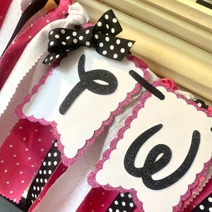 May include: A decorative banner with white, pink, and black polka dot ribbons. The banner features white scalloped pennants with the letters "Y" and "W" in black glitter. A black bow with white polka dots is attached.