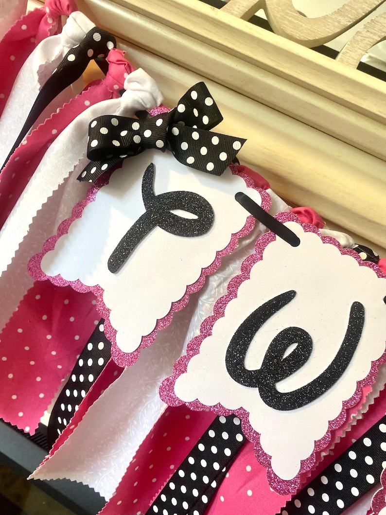 May include: A decorative banner with white, pink, and black polka dot ribbons. The banner features white scalloped pennants with the letters "Y" and "W" in black glitter. A black bow with white polka dots is attached.