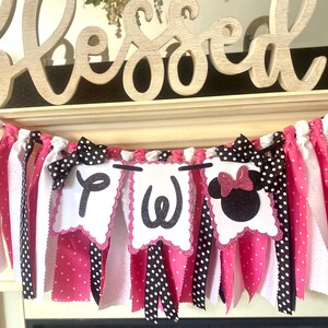 May include: A decorative banner with pink, white, and black fabric strips. The banner features three white pennants with the letters "P", "W", and a Minnie Mouse head. Black polka dot bows and the word "Blessed" are also visible.