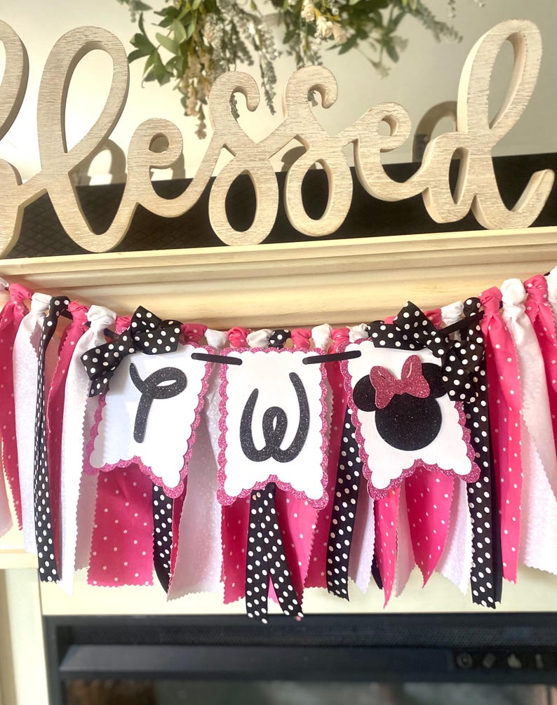 May include: A decorative banner with pink, white, and black fabric strips. The banner features three white pennants with the letters "P", "W", and a Minnie Mouse head. Black polka dot bows and the word "Blessed" are also visible.