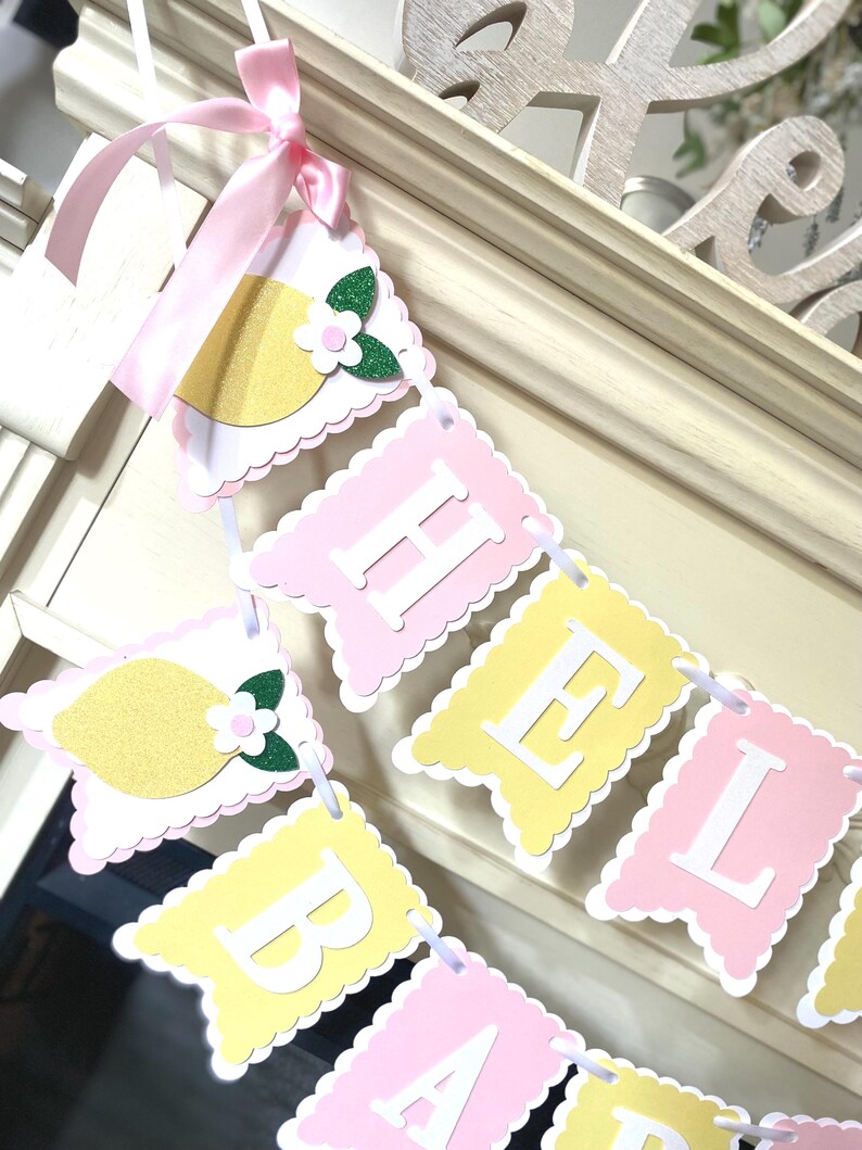 May include: A decorative banner with alternating pink and yellow pennants spelling "HELLO BABY". Each pennant has a scalloped edge and is strung together with white ribbon. Lemon and flower cutouts adorn the banner ends, tied with a pink ribbon.