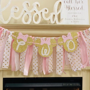 May include: A pink and gold banner with Minnie Mouse-shaped cutouts spelling "ONE". The banner has pink bows, ribbons, and polka dot fabric. The word "Blessed" is above the banner, and a sign reads "Her Children Rise Up and Call Her Blessed."