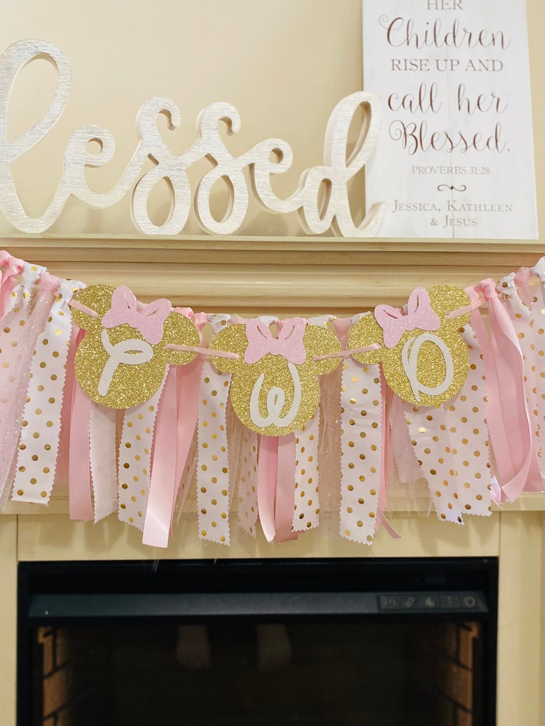 May include: A pink and gold banner with Minnie Mouse-shaped cutouts spelling "ONE". The banner has pink bows, ribbons, and polka dot fabric. The word "Blessed" is above the banner, and a sign reads "Her Children Rise Up and Call Her Blessed."