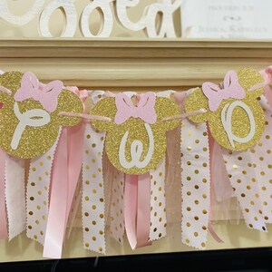 May include: A festive banner with gold glitter Minnie Mouse head shapes spelling "One". Each head has a pink bow. The banner is strung with pink and white polka dot fabric and pink ribbons, perfect for a birthday celebration.