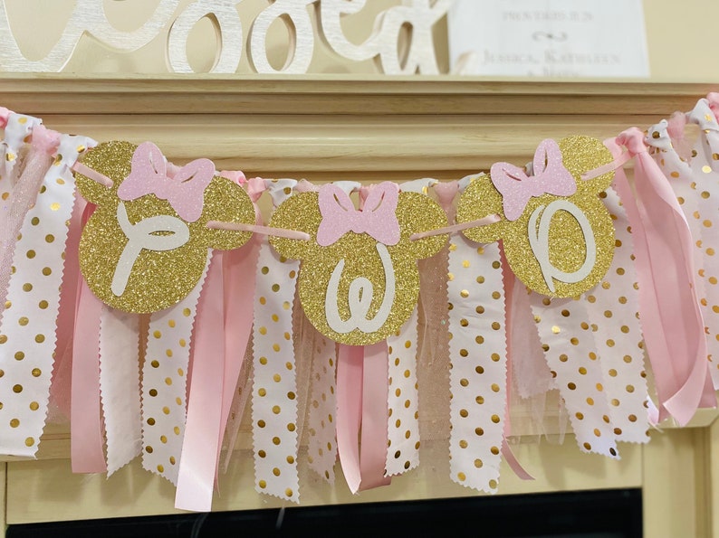 May include: A festive banner with gold glitter Minnie Mouse head shapes spelling "One". Each head has a pink bow. The banner is strung with pink and white polka dot fabric and pink ribbons, perfect for a birthday celebration.