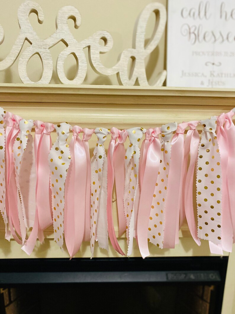 May include: A pink and gold polka dot ribbon garland. The garland features alternating ribbons of solid pink, white with gold polka dots, and white with a scalloped edge. The garland is hanging in front of a fireplace.