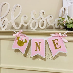 Puede incluir: Bandera rosa y dorada con la palabra "ONE" y una cabeza de Minnie Mouse. La bandera est&aacute; decorada con lazos rosas y est&aacute; colgada de una repisa de chimenea blanca.