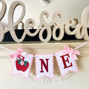May include: A white and pink banner with the word "ONE" in red glitter letters. The banner has a red glitter apple with a green stem and a pink flower on the left side. The banner is decorated with pink bows.