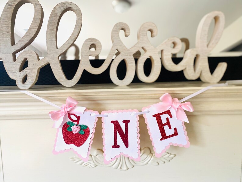 May include: A white and pink banner with the word "ONE" in red glitter letters. The banner has a red glitter apple with a green stem and a pink flower on the left side. The banner is decorated with pink bows.