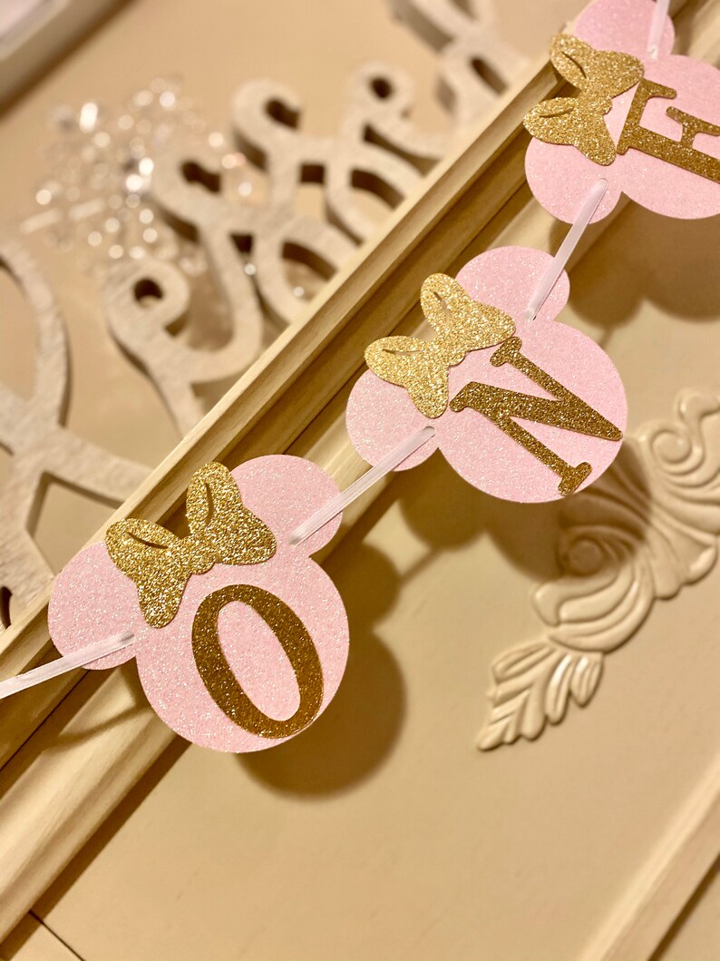 Op de afbeelding: Roze en gouden glitterbanner met het woord "EEN" in gouden letters en een gouden strik op elke letter. De banner is perfect voor een eerste verjaardagsparty.