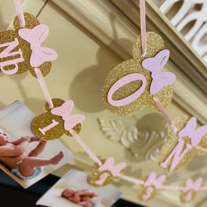 May include: A pink and gold glitter banner with the letters "ONE" and a pink bow on each letter. The banner is strung with pink ribbon and has a photo of a baby on each letter.