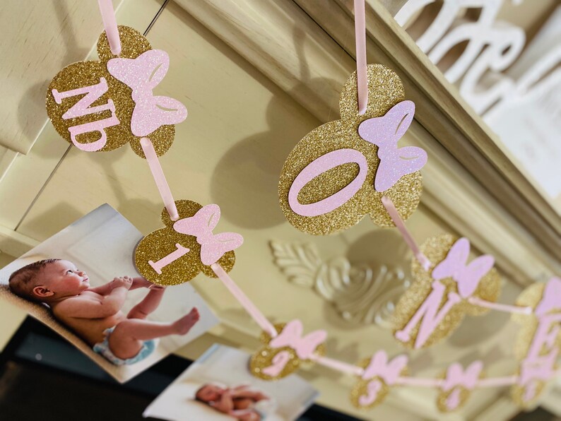 May include: A pink and gold glitter banner with the letters "ONE" and a pink bow on each letter. The banner is strung with pink ribbon and has a photo of a baby on each letter.