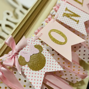 May include: A pink and gold banner with the word "ONE" and Minnie Mouse-themed decorations. The banner features pennant flags with polka dots, gold glitter accents, and pink ribbons. The banner is designed for a first birthday celebration.
