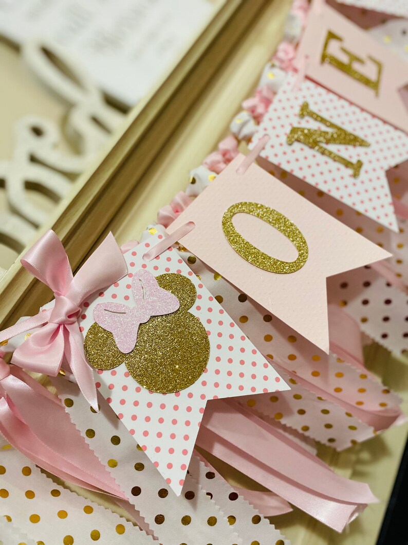 May include: A pink and gold banner with the word "ONE" and Minnie Mouse-themed decorations. The banner features pennant flags with polka dots, gold glitter accents, and pink ribbons. The banner is designed for a first birthday celebration.