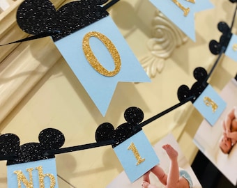 May include: A blue and black Mickey Mouse themed banner with gold glitter letters spelling "One" and a photo of a baby. The banner is strung with black ribbon and has black Mickey Mouse ears between each letter.