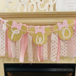 May include: A festive banner with gold glitter Minnie Mouse head cutouts spelling "ONE" in white. Pink bows and ribbons hang below the cutouts, with gold polka dot fabric. The banner is a decorative element, likely for a first birthday celebration.