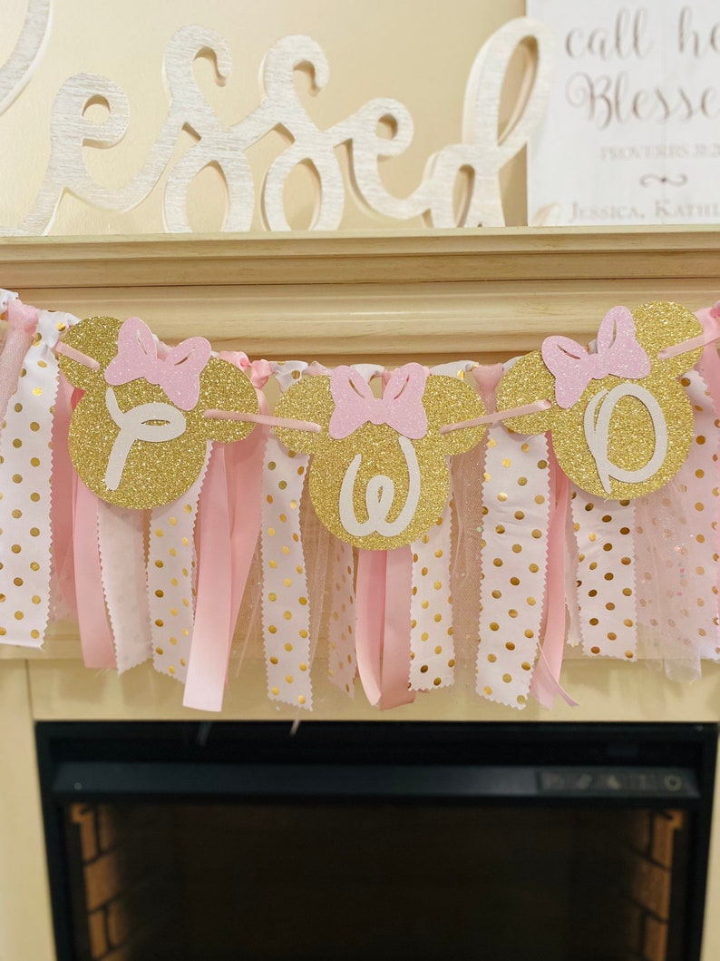 May include: A festive banner with gold glitter Minnie Mouse head cutouts spelling "ONE" in white. Pink bows and ribbons hang below the cutouts, with gold polka dot fabric. The banner is a decorative element, likely for a first birthday celebration.