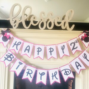 May include: A pink and white banner with black letters that reads "Happy 2nd Birthday". The banner is decorated with pink glitter and has a scalloped edge. The banner is hanging from a wooden sign that reads "blessed".