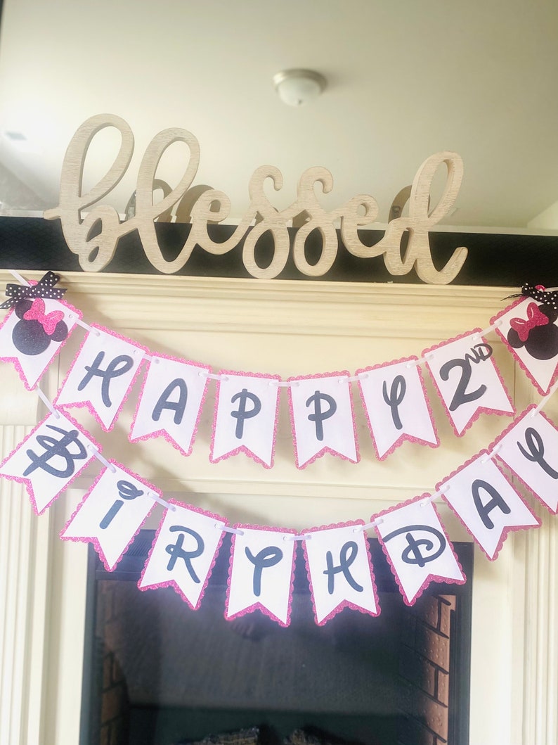 May include: A pink and white banner with black letters that reads "Happy 2nd Birthday". The banner is decorated with pink glitter and has a scalloped edge. The banner is hanging from a wooden sign that reads "blessed".
