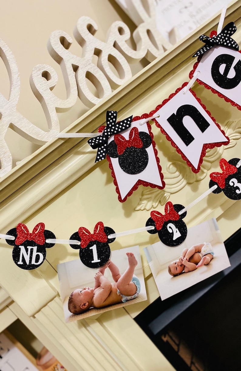 May include: A red and black Minnie Mouse themed first birthday banner with black glitter numbers and red bows. The banner includes photos of a baby at different ages.