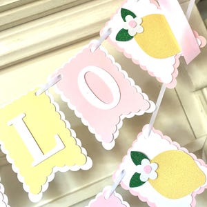 May include: A decorative banner with pink and yellow pennants. The banner spells out "LOVEY" in white letters. Each pennant has a scalloped edge and is decorated with a lemon and flower design. A pink ribbon is attached at the top.