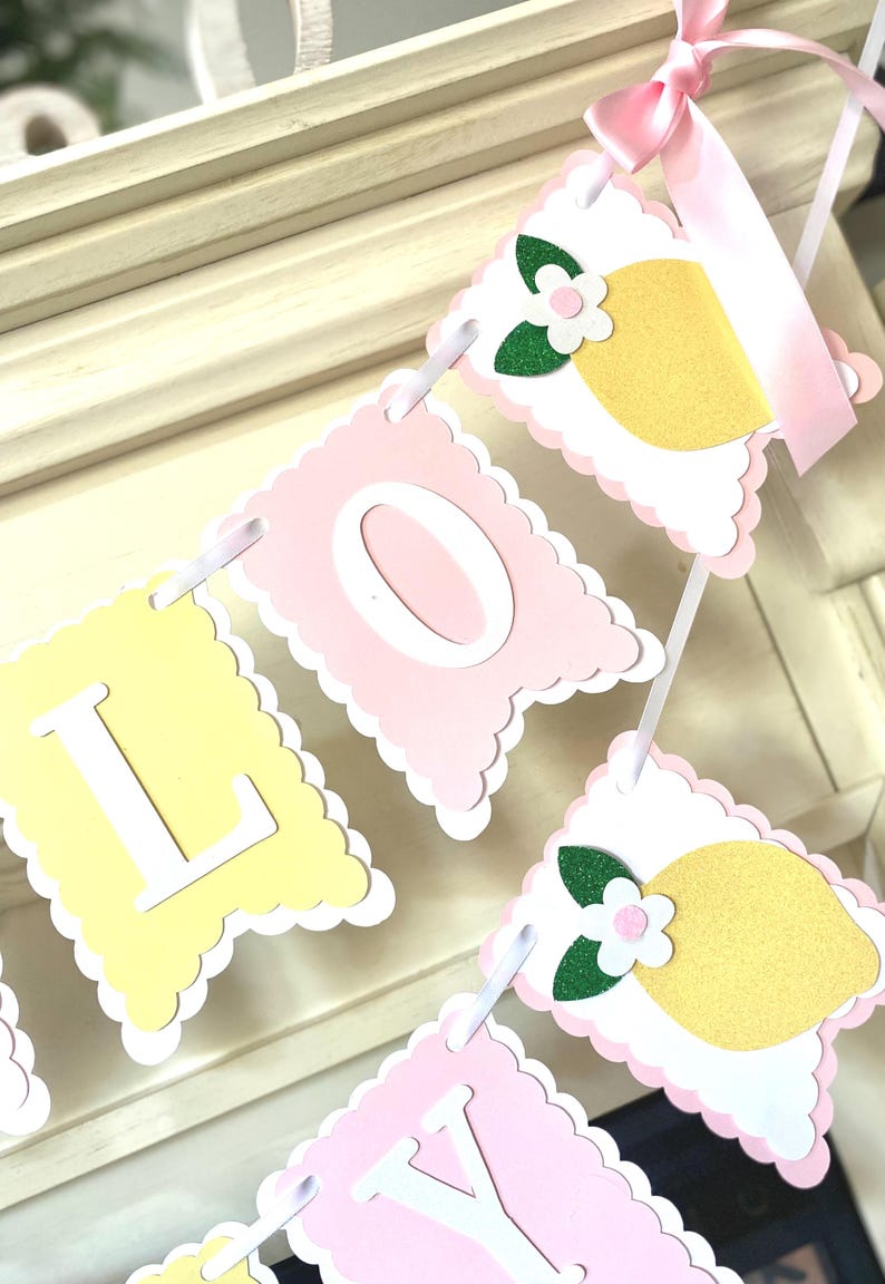 May include: A decorative banner with pink and yellow pennants. The banner spells out "LOVEY" in white letters. Each pennant has a scalloped edge and is decorated with a lemon and flower design. A pink ribbon is attached at the top.