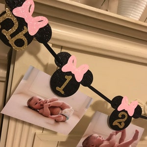 May include: A black and gold glitter banner with Minnie Mouse-themed cutouts, pink bows, and numbers 0-3. The banner holds photos of a baby. A decorative piece for a baby's birthday.