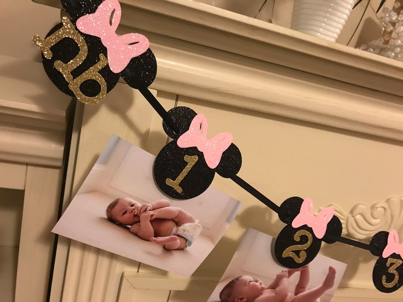 May include: A black and gold glitter banner with Minnie Mouse-themed cutouts, pink bows, and numbers 0-3. The banner holds photos of a baby. A decorative piece for a baby's birthday.