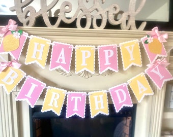 May include: A pink and yellow banner with the words "Happy Birthday" in white letters. The banner has scalloped edges and is decorated with small pink bows and yellow lemons with green leaves.