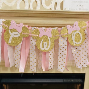 May include: A gold glitter banner with pink bows and the word "One" in white. The banner is strung with pink ribbon and white fabric with gold polka dots. The banner is likely for a birthday celebration.