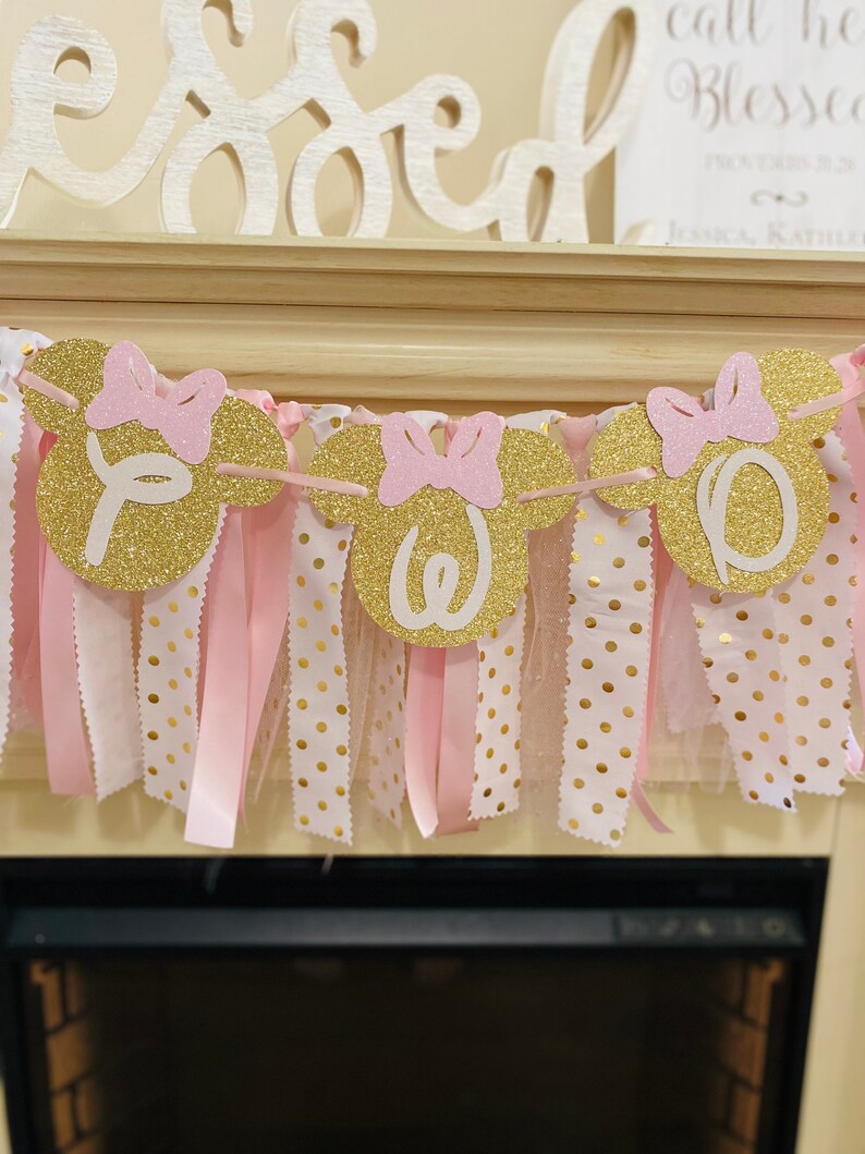 May include: A gold glitter banner with pink bows and the word "One" in white. The banner is strung with pink ribbon and white fabric with gold polka dots. The banner is likely for a birthday celebration.