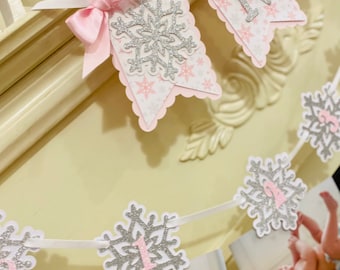 May include: A pink and silver snowflake banner with photos of a baby. The banner has scalloped edges and a pink bow. The banner spells out "ONE" with silver glitter letters and snowflake cutouts.