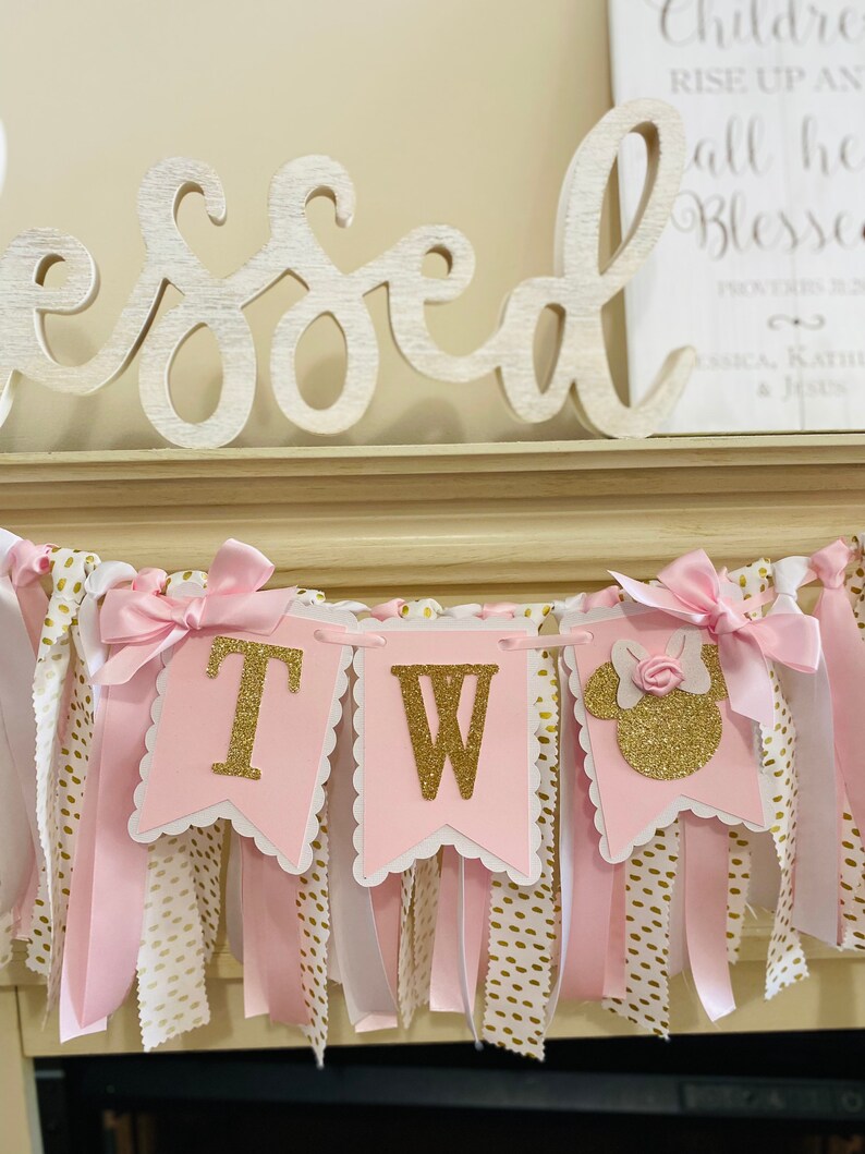 May include: Pink and gold banner with the letters "TW" and a Minnie Mouse silhouette. The banner has pink and white ribbons with gold polka dots and is decorated with pink bows. The word "Blessed" is in the background.