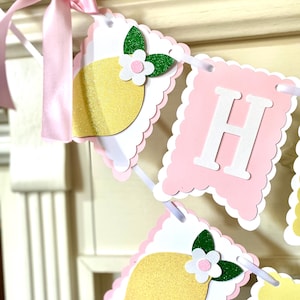 May include: A decorative banner with pink and white scalloped edges. The banner features lemon and letter cutouts. The lemons are yellow with glitter, and the letters are white. A pink ribbon is tied to the banner.