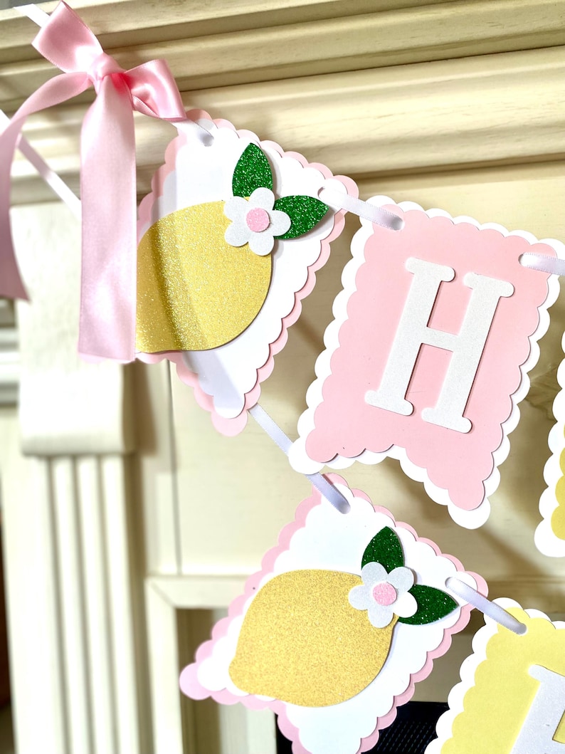 May include: A decorative banner with pink and white scalloped edges. The banner features lemon and letter cutouts. The lemons are yellow with glitter, and the letters are white. A pink ribbon is tied to the banner.