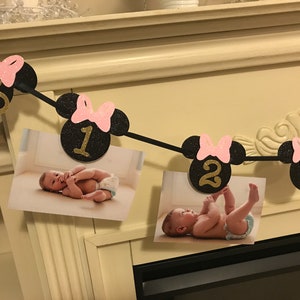 May include: A black and pink Minnie Mouse-themed photo banner with the numbers "NB", "1", "2", and "3" in gold glitter. The banner holds photos of a baby. The banner is strung across a white mantel.