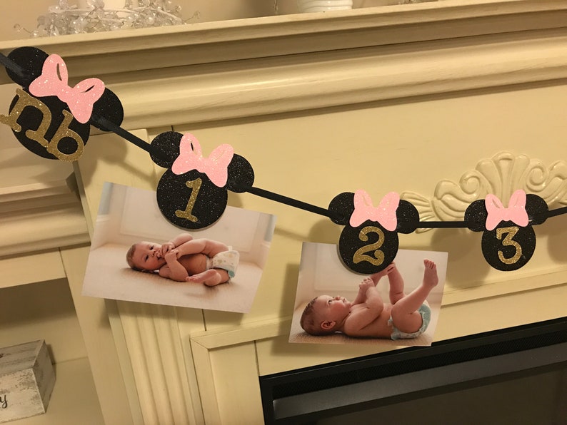 May include: A black and pink Minnie Mouse-themed photo banner with the numbers "NB", "1", "2", and "3" in gold glitter. The banner holds photos of a baby. The banner is strung across a white mantel.