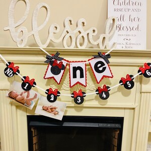 May include: A red and black Minnie Mouse themed banner with the word "one" in white letters. The banner is decorated with red and black polka dots and has a white string for hanging. The banner is hanging above a fireplace with a white mantle. The text "Blessed" is written on a white sign above the banner.