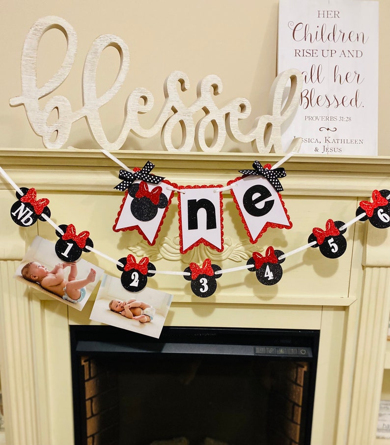 May include: A red and black Minnie Mouse themed banner with the word "one" in white letters. The banner is decorated with red and black polka dots and has a white string for hanging. The banner is hanging above a fireplace with a white mantle. The text "Blessed" is written on a white sign above the banner.