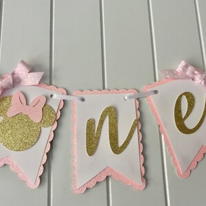 Puede incluir: Pancarta de "uno" con purpurina rosa y dorada con una cabeza de Minnie Mouse de purpurina rosa y dorada en el lado izquierdo. La pancarta est&aacute; enhebrada con cinta blanca y tiene lazos de lunares rosas en cada extremo.