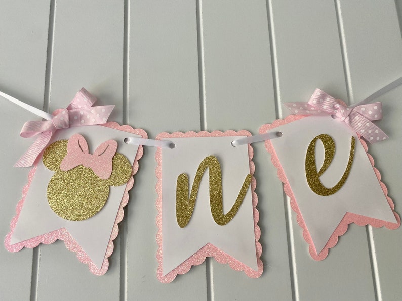 Puede incluir: Pancarta de "uno" con purpurina rosa y dorada con una cabeza de Minnie Mouse de purpurina rosa y dorada en el lado izquierdo. La pancarta est&aacute; enhebrada con cinta blanca y tiene lazos de lunares rosas en cada extremo.