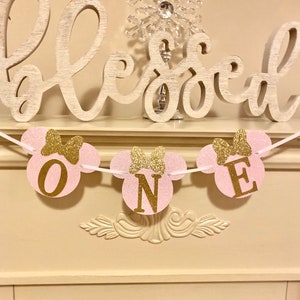 Op de afbeelding: Een roze en gouden glitter Minnie Mouse-themabanner met het woord "ONE" in gouden letters. De banner hangt aan een wit touw en is versierd met gouden strikken. De banner hangt voor een witte open haard met een houten bord waarop "Blessed" staat.