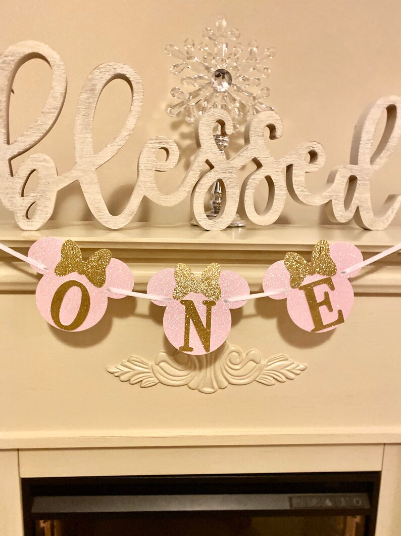 Op de afbeelding: Een roze en gouden glitter Minnie Mouse-themabanner met het woord "ONE" in gouden letters. De banner hangt aan een wit touw en is versierd met gouden strikken. De banner hangt voor een witte open haard met een houten bord waarop "Blessed" staat.