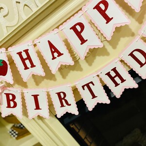 May include: A pink and white banner with red glitter letters that reads "Happy Birthday". The banner has a scalloped edge and is strung together with white string. There is a red apple with a green stem and a pink bow on the left side of the banner.