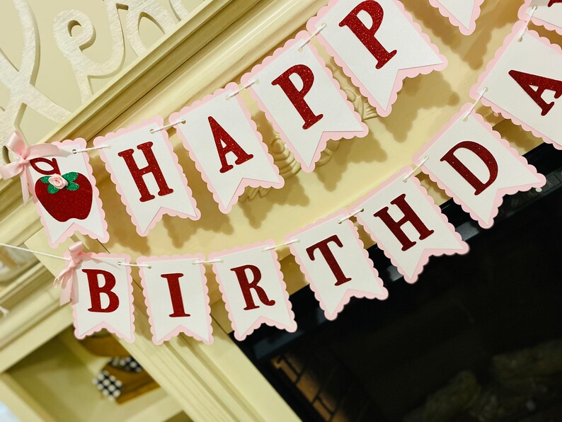 May include: A pink and white banner with red glitter letters that reads "Happy Birthday". The banner has a scalloped edge and is strung together with white string. There is a red apple with a green stem and a pink bow on the left side of the banner.