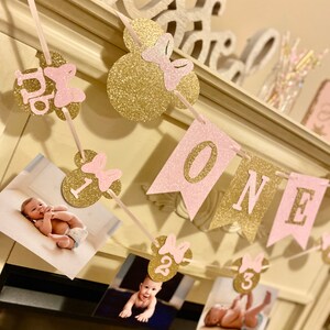 Puede incluir: Una pancarta de brillo rosa y dorado con la palabra "ONE" y orejas de Mickey Mouse con lazos. La pancarta est&aacute; decorada con fotos de una ni&ntilde;a peque&ntilde;a a diferentes edades.