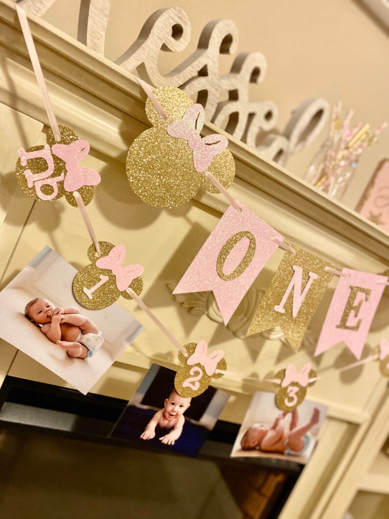 Puede incluir: Una pancarta de brillo rosa y dorado con la palabra "ONE" y orejas de Mickey Mouse con lazos. La pancarta est&aacute; decorada con fotos de una ni&ntilde;a peque&ntilde;a a diferentes edades.