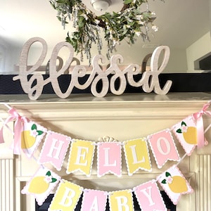 May include: A decorative mantel arrangement featuring a wooden "Blessed" sign, a floral wreath, and two banners. The banners read "HELLO BABY" with lemon and flower accents, perfect for a baby shower or nursery decor.